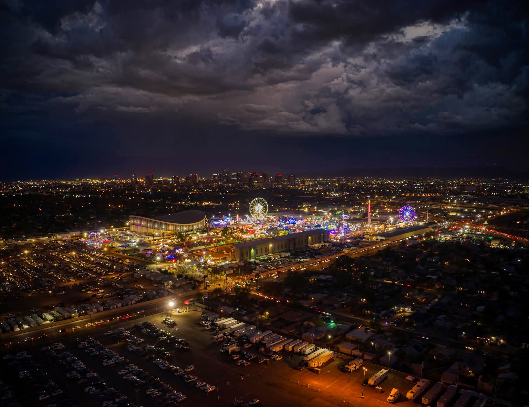 Wide aerial night photography of the Arizona State Fair in Phoenix, featuring glowing carnival rides, a lit Ferris wheel, and the city skyline under a dramatic cloudy night sky.