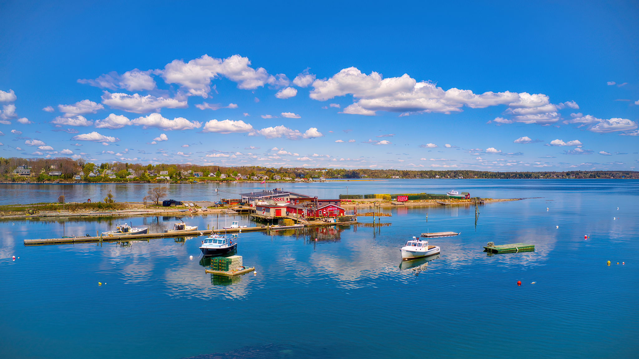 Wide aerial drone photography of Cooks Lobster House and Garrison Cove harbor in Bailey Island, Maine, featuring calm blue water and boats at the docks under a clear sky.