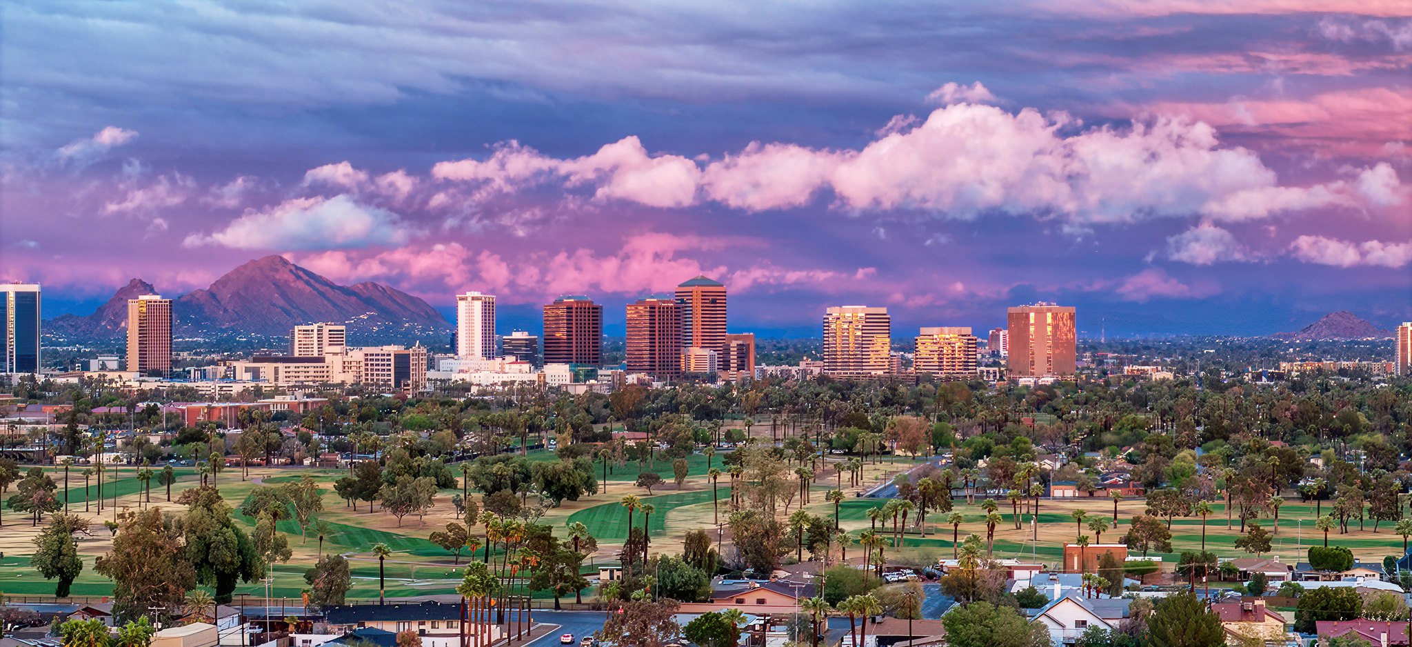 Wide panoramic aerial view of the Phoenix city skyline and lush green urban parks under a vibrant purple and pink sunset sky.
