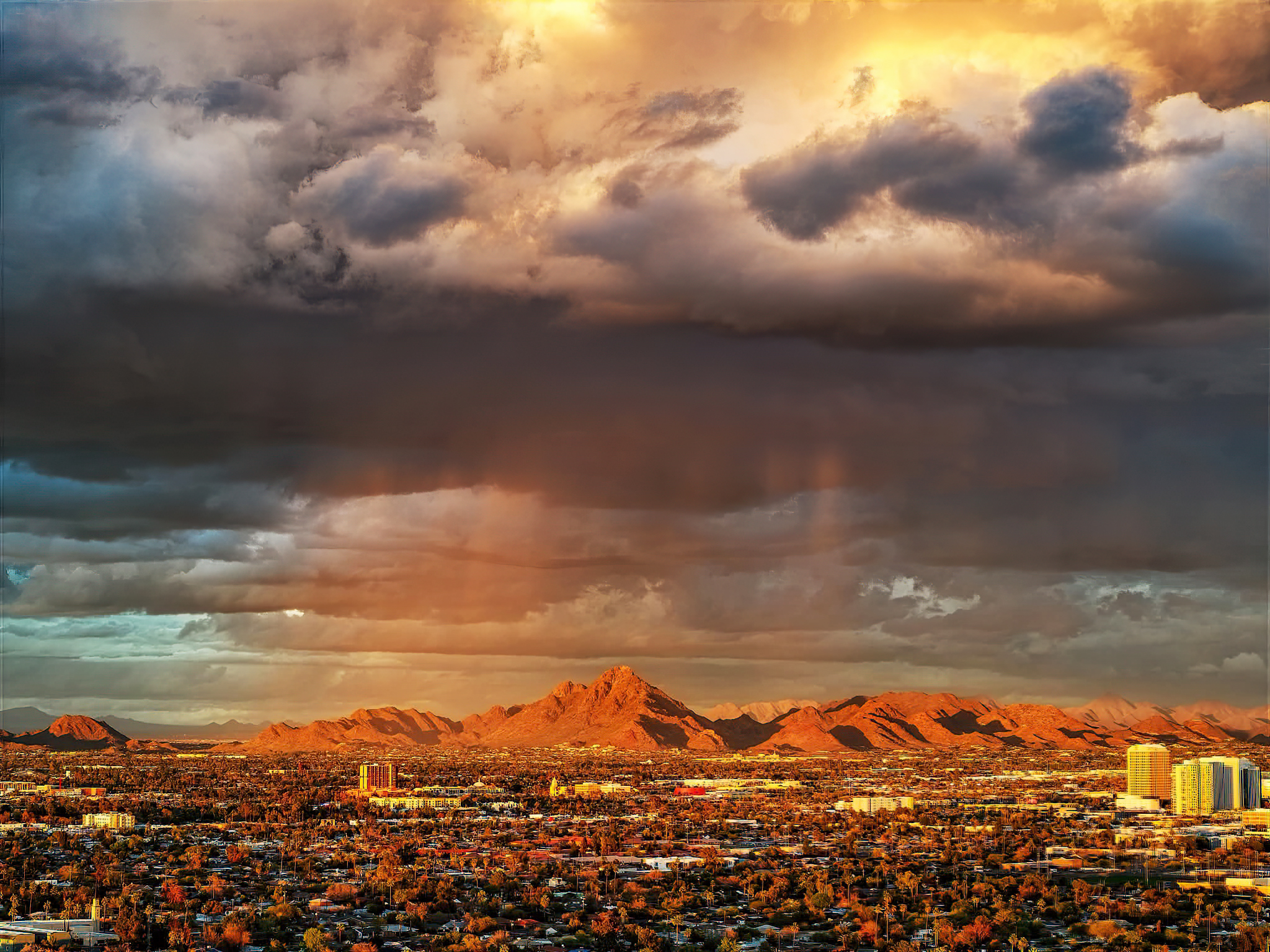 Aerial view of Arizona mountain peaks glowing in golden sunlight under a dramatic storm-cloud sky.